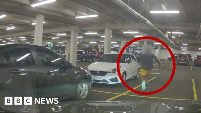 A man in blue sneakers is circled in red, running through a car park.