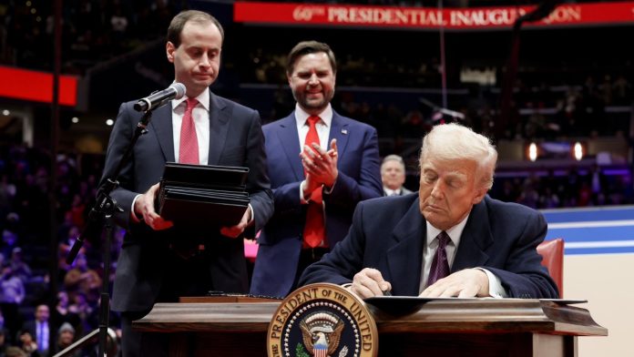 US President Donald Trump signs a document at a desk as aides stand behind him