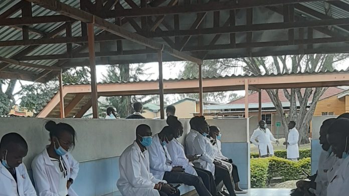 Health workers in white coats sit in a row along a bench at a health facility